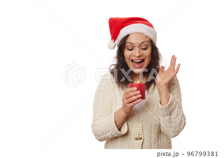 Hispanic surprised beautiful woman in Santa Claus hat and warm beige sweater, looking at a lit red candle on white background. Christmas atmosphere. Free advertising space for your promotional text Hispanic surprised beautiful woman in Santa Claus hat and warm beige sweater, looking at a lit red candle on white background. Christmas atmosphere. Free advertising space for your promotional text 96799381