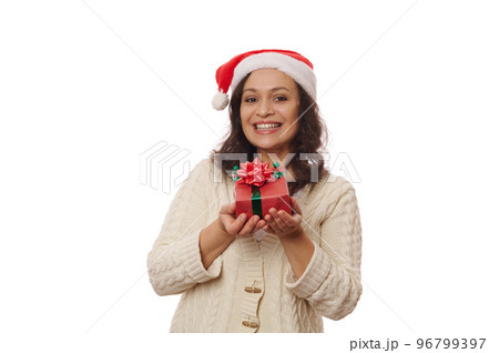 Multi-ethnic brunette, a happy middle-aged woman in Santa hat and beige sweater, showing a Christmas present in red wrapping paper and bow, cutely smiling, looking at camera, against white background 96799397