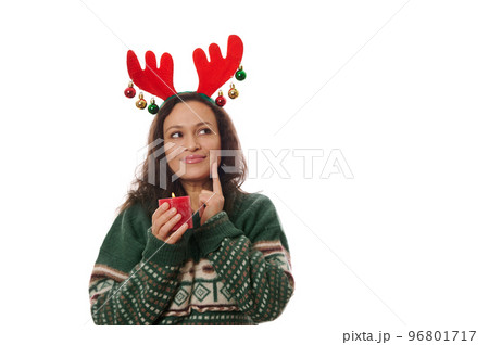 Isolated on white background, charming delightful multi-ethnic woman in warm Xmas patterned green sweater and deer antler hoop, holding a red lit Christmas candle and smiling looking up at copy space 96801717