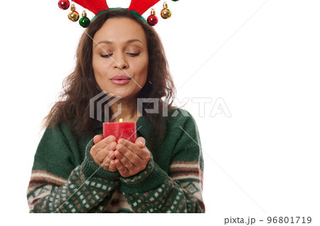 Attractive Latin American brunette woman, wearing warm wool green sweater and deer antler hoop, making cherished wish while blowing out a red candle, isolated on white background with free ad space. Attractive Latin American brunette woman, wearing warm wool green sweater and deer antler hoop, making cherished wish while blowing out a red candle, isolated on white background with free ad space. 96801719