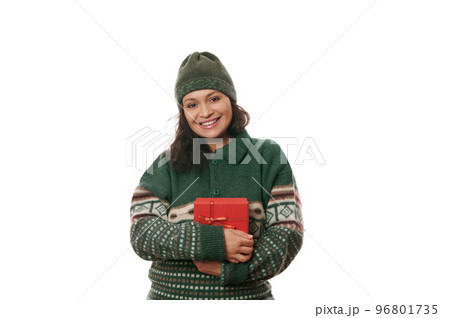 Merry Christmas. Portrait of a beautiful multi-ethnic woman in a cozy knitted green Xmas patterned sweater and wool green hat, holding gift box, smiling looking at camera, isolated on white background 96801735
