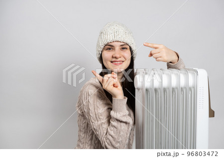 A woman basks near an electric oil heater and points her fingers to the side in place for text. Methods of heating an apartment or house. 96803472