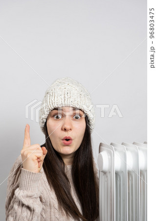 A woman in a hat getting warm near the oil heater and looks and points up with her finger. 96803475