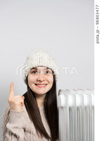 A woman in a hat getting warm near the oil heater and looks and points up with her finger. 96803477