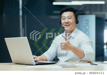 A young male student sits in an office, co-working space, library at a desk in a headset. learns online remotely from a laptop. He looks at the camera, points a super finger, smiles A young male student sits in an office, co-working space, library at a desk in a headset. learns online remotely from a laptop. He looks at the camera, points a super finger, smiles 96804151