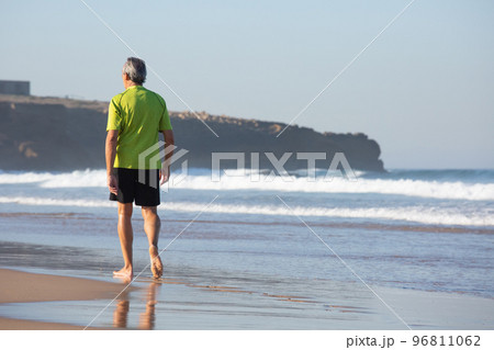 Back view of aged man walking barefoot along seashore 96811062