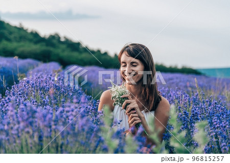 Close up portrait of happy young brunette woman in white dress on blooming fragrant lavender fields with endless rows. Warm sunset light. Bushes of lavender purple aromatic flowers on lavender fields. Close up portrait of happy young brunette woman in white dress on blooming fragrant lavender fields with endless rows. Warm sunset light. Bushes of lavender purple aromatic flowers on lavender fields. 96815257