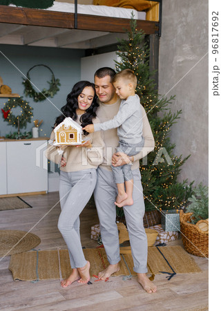 A stylish young family with a small child is standing in their kitchen, decorated for Christmas, and admiring a gingerbread house prepared with their own hands 96817692