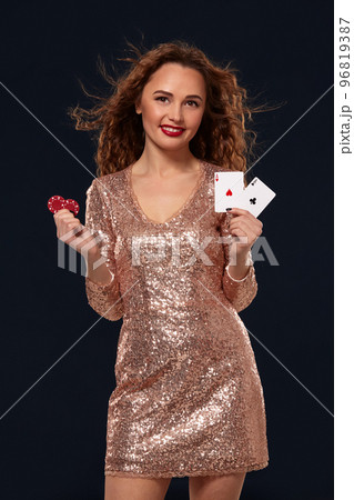 Brown-haired attractive caucasian young woman in golden cocktail dress at casino holds pair of aces and chips in her hands 96819387