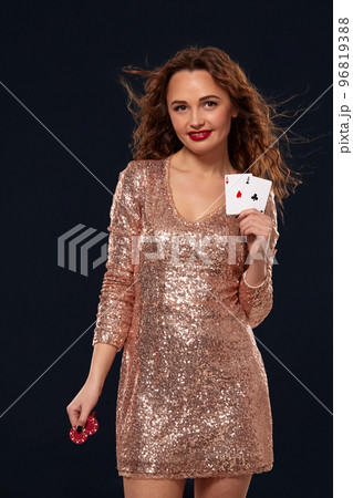 Brown-haired attractive caucasian young woman in golden cocktail dress at casino holds pair of aces and chips in her hands 96819388