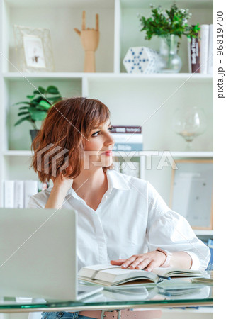 Woman in the office at the computer. She is dressed in a white shirt and blue jeans sitting at a table on which is a laptop and a notepad. Works in the office, bright workplace. 96819700