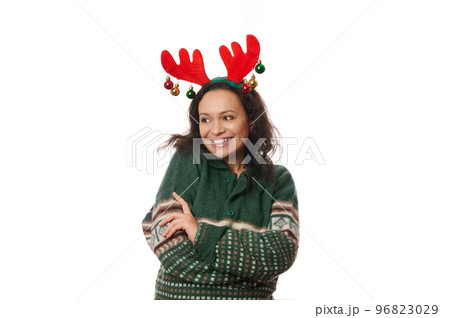 Multi-ethnic joyful attractive woman, wearing deer antler hoop and warm green sweater with Christmas pattern, hugging herself, laughing, looking aside at copy advertising space on white background Multi-ethnic joyful attractive woman, wearing deer antler hoop and warm green sweater with Christmas pattern, hugging herself, laughing, looking aside at copy advertising space on white background 96823029