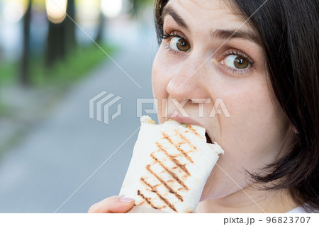 Brunette woman overeats shawarma on a city street. Street fast food pita roll with meat and vegetables. Place for text. Brunette woman overeats shawarma on a city street. Street fast food pita roll with meat and vegetables. Place for text. 96823707