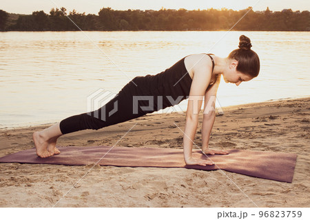 A woman stands in a plank pose on the river bank on the beach at sunset in summer. Yoga or fitness classes in nature. 96823759