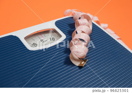 Floor blue scales and centimeter measuring tape on an orange background, top view. The concept of weight loss and weight control Floor blue scales and centimeter measuring tape on an orange background, top view. The concept of weight loss and weight control 96825301