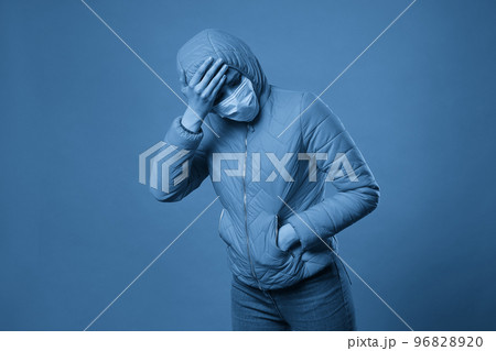 woman wearing hood in a protective face mask and coat over blue background. Model girl in medical mask and spring outfit, studio shot with copy space for your text. Anti-flu, anti-epidemic woman wearing hood in a protective face mask and coat over blue background. Model girl in medical mask and spring outfit, studio shot with copy space for your text. Anti-flu, anti-epidemic 96828920