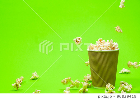 Popcorn is poured into a paper brown glass on a green background with place for text. Popcorn is poured into a paper brown glass on a green background with place for text. 96829419