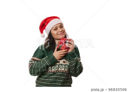 Attractive multi-ethnic woman in Santa hat and warm green sweater, holding Christmas present near her face, smiling cutely looking at camera, isolated over white background with free advertising space 96830506