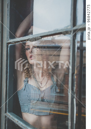 Beautiful attractive woman with curly hait and grey top staying in the window and looking 96842775