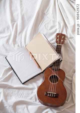 Ukulele guitar and open notebook on white bed. Cozy atmosphere for writing music. Ukulele guitar and open notebook on white bed. Cozy atmosphere for writing music. 96842972