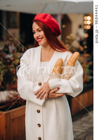 Portrait of a pretty woman in a red beret and a white cardigan with baguettes in her hands strolling through the autumn city Portrait of a pretty woman in a red beret and a white cardigan with baguettes in her hands strolling through the autumn city 96844441