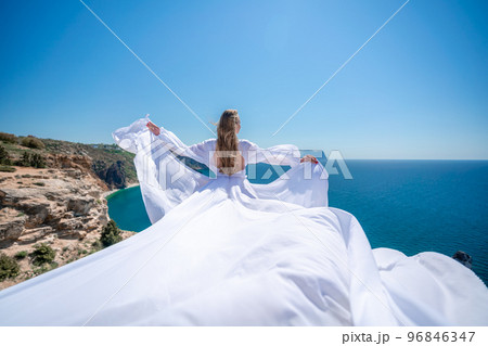 Blonde with long hair on a sunny seashore in a white flowing dress, rear view, silk fabric waving in the wind. Against the backdrop of the blue sky and mountains on the seashore. Blonde with long hair on a sunny seashore in a white flowing dress, rear view, silk fabric waving in the wind. Against the backdrop of the blue sky and mountains on the seashore. 96846347