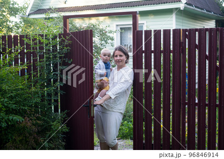 Curious woman with a baby opening the gate of fence and looking at camera Curious woman with a baby opening the gate of fence and looking at camera 96846914