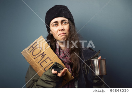 A beggar suffering caucasian woman holds a cardboard sign with the inscription homeless and begging with alms cup. Dark background. The concept of helping vagrants 96848030
