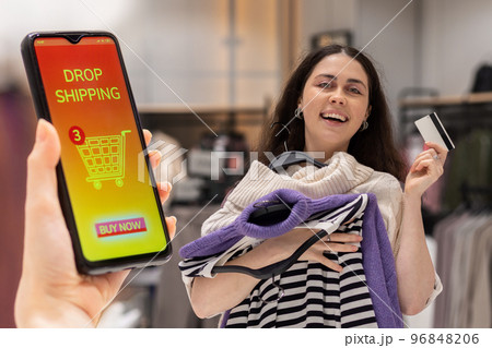 Happy young woman holds hangers with clothes and bank card. Close up of female hand holding smartphone with online store at screen. Concept of online shopping and drop shipping 96848206