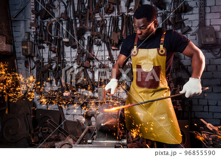 handsome african american blacksmith male worker working in workshop,wearing leather apron and protective glasses with gloves.concept save work 96855590