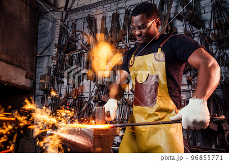 handsome african americam man forging steel next to furnace in dark workshop. small business comcept handsome african americam man forging steel next to furnace in dark workshop. small business comcept 96855771