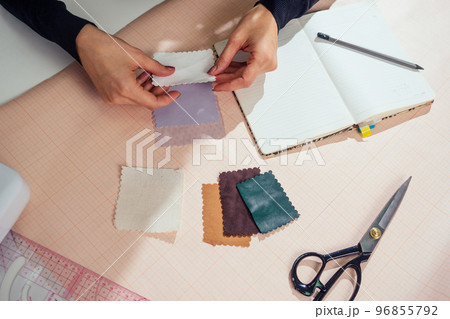 beautiful seamstress with long hair writes in a notebook. the tailor creates a collection of outfits. young woman designer clothes working with leather beautiful seamstress with long hair writes in a notebook. the tailor creates a collection of outfits. young woman designer clothes working with leather 96855792