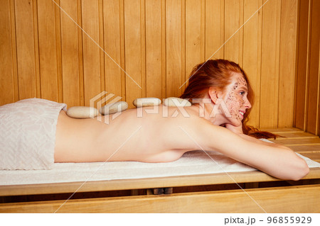 redhaired ginger young woman peeling face with brown sugar in traditional russian banya.steaming and self-care in finnish sauna taking stones massaging 96855929