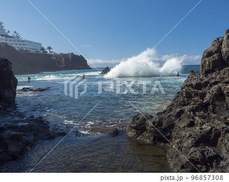 Big waves splash against natural rock swimming pool Charco de Isla Cangrejo in the Atlantic Ocean in Los Gigantes, Tenerife, Canary islands, Spain. Sunny winter day. Big waves splash against natural rock swimming pool Charco de Isla Cangrejo in the Atlantic Ocean in Los Gigantes, Tenerife, Canary islands, Spain. Sunny winter day. 96857308