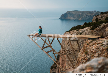 A girl sits on a wooden springboard for jumping with a rope. In a dark green dress with her hands in the air. A girl sits on a wooden springboard for jumping with a rope. In a dark green dress with her hands in the air. 96860491