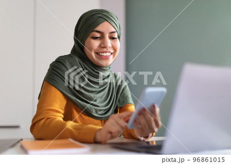 Muslim Woman Shopping Online With Smartphone While Sitting At Desk In Office Muslim Woman Shopping Online With Smartphone While Sitting At Desk In Office 96861015
