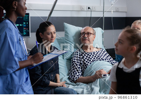 African american female physician filling out medical interventions, surgery record for senior palliative care patient. Sick elderly man family listening medication instructions from young nurse. 96861310
