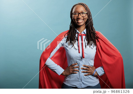 Proud and happy young superhero woman posing brave for camera while standing on blue background. Strong and selfless justice defender wearing mighty hero red cloak while standing with hands on hip. Proud and happy young superhero woman posing brave for camera while standing on blue background. Strong and selfless justice defender wearing mighty hero red cloak while standing with hands on hip. 96861326