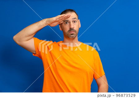 A man in greeting pose and serious face. A hispanic man with a beard wearing orange t-shirt isolated on blue background 96873529