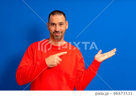 Cheerful bearded man pointing finger holding something on palm. Man in his 40s wearing a red pullover, isolated on a blue background. Sale, advertisement, promotion concept. Close up. 96873531