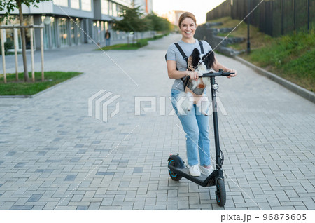 A woman rides an electric scooter with a dog in a backpack. Pappilion Spaniel Continental in a sling. 96873605