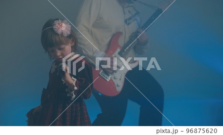 Little girl on stage in vintage dress, she sings into microphone and dances, her father plays an electric guitar. Color music is shining and smoke is billowing. Family time, creativity and hobbies Little girl on stage in vintage dress, she sings into microphone and dances, her father plays an electric guitar. Color music is shining and smoke is billowing. Family time, creativity and hobbies 96875620