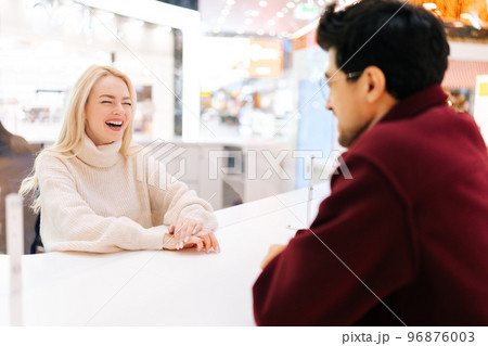 Back view from shoulder of unrecognizable young man chatting through glass partition with cheerful blonde woman standing in hall of shopping mall. Concept of lifestyle social distancing and work. Back view from shoulder of unrecognizable young man chatting through glass partition with cheerful blonde woman standing in hall of shopping mall. Concept of lifestyle social distancing and work. 96876003
