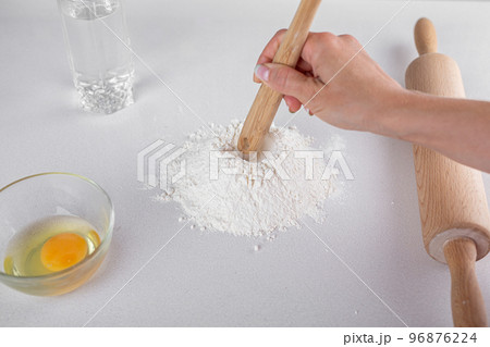 Top view of mound the flour, egg in glass, wooden spoon, water, rolling pin on white table. Dough Top view of mound the flour, egg in glass, wooden spoon, water, rolling pin on white table. Dough 96876224