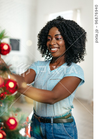 Beautiful young african woman decorating Christmas tree at home Beautiful young african woman decorating Christmas tree at home 96876940