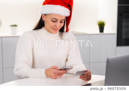 An excited woman in a red Santa Claus hat uses a credit card and mobile phone to order products for a Christmas party.  96878509
