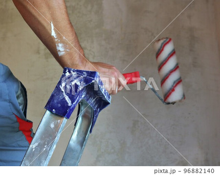 painter's hands smeared in paint with a red and white roller hold the top of an iron stepladder in a room being renovated, the concept of home renovation work with wall painting and wallpapering painter's hands smeared in paint with a red and white roller hold the top of an iron stepladder in a room being renovated, the concept of home renovation work with wall painting and wallpapering 96882140
