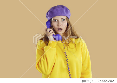 Stunned puzzled woman in purple hat and yellow sweatshirt calling phone on beige background. 96883282