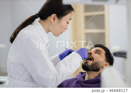 Asian dentist examines the oral cavity of young bearded handsome man Asian dentist examines the oral cavity of young bearded handsome man 96888527