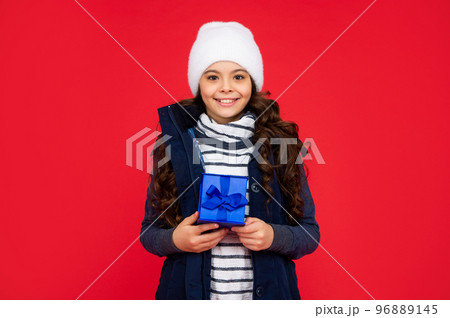 happy child in puffer jacket and hat hold box. kid with present. teen girl on red background happy child in puffer jacket and hat hold box. kid with present. teen girl on red background 96889145